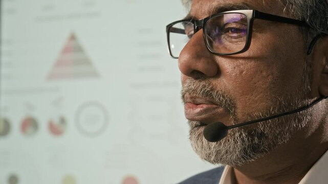 Close-up shot of middle-aged biracial man with beard and glasses talking into microphone while delivering presentation on stage
