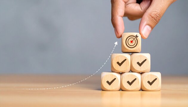 A conceptual digital illustration representing goal setting, achievement, and success. A hand places a wooden cube with a target and arrow symbol on top of a pyramid made of five other wooden cubes, e