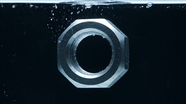 A close-up shot of a silver metal nut submerged in dark water with small bubbles around it.
