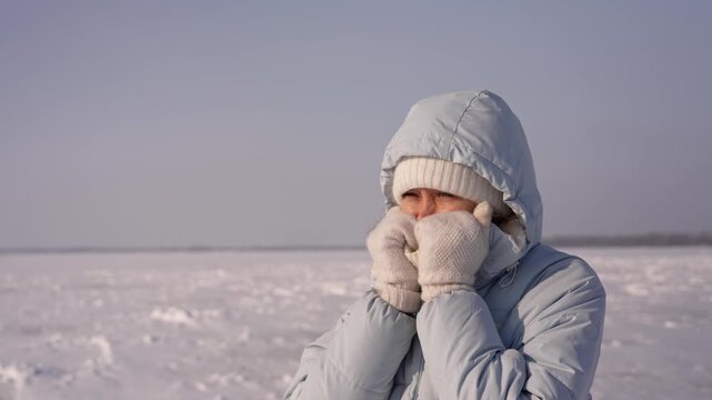 Freezing woman in a warm jacket trying to keep warm with her hands on a sunny but very cold day, standing on a frozen river covered with snow during an arctic winter expedition
