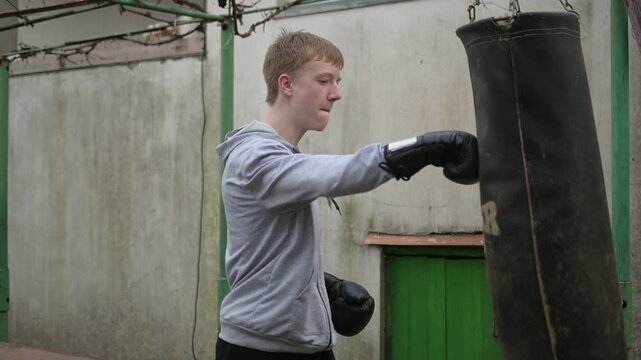 Young Man in Grey Hoodie Training with Heavy Punching Bag Outdoors
