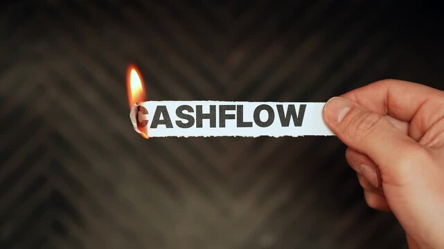 Close-up hand burns a paper label reading &ldquo;CASHFLOW&rdquo; with a lighter flame on dark background&mdash;finance stress, liquidity problems, budgeting crisis concept, copy space.