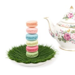 Stack of colorful macarons on a bed of green grass with a floral teapot nearby.