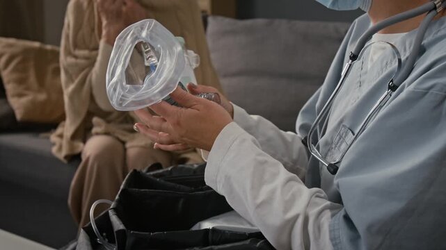 Tilt up view of young female health worker with face mask and stethoscope around neck opening first aid bag and showing medical devices and pill bottle to sick elderly lady