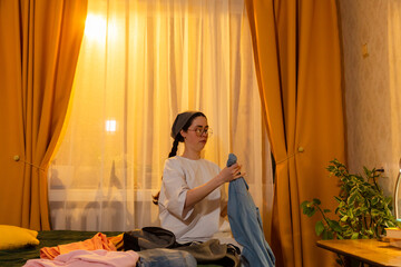 Obraz premium Young Caucasian woman tidying her room while folding clothes indoors. Sunset light fills the space. Clean home theme. Low angle view