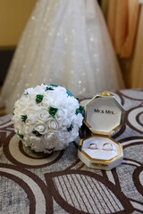 White rose bouquet and ring box with Mr. & Mrs. on a bed against a wedding dress background.