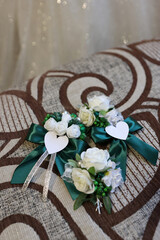 Wedding boutonnieres with white flowers and green ribbons shaped with hearts on a bed.