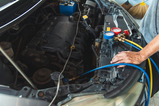 A close-up view of an auto mechanic's hands using a manifold gauge set to test and recharge the air conditioning system in a car's engine bay.