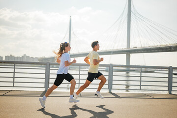Two people run along a riverside path under a bridge on a sunny day