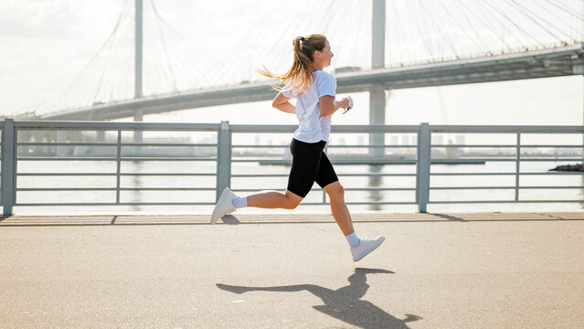 Woman runs along a path beside water, with a bridge in view during daytime