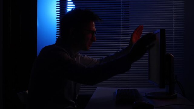 Man in silhouette reacting with frustration while working on a computer in a dark room late at night, illuminated by the screen and a blue light from outside, depicting stress and cyber issues