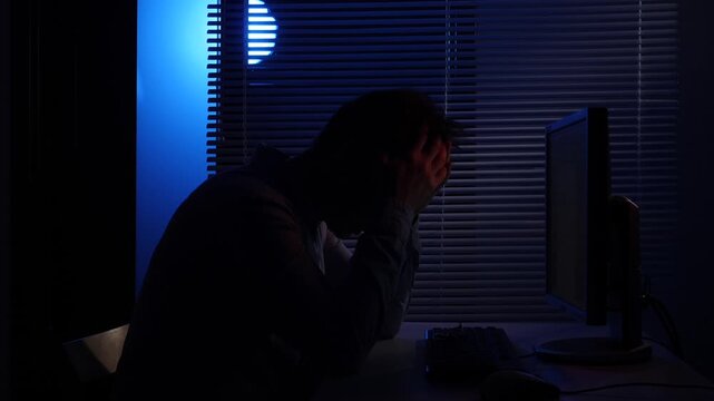 Silhouette of a frustrated man sitting at a desk in a dark room, hitting his head, with hands covering his head, illuminated by a computer screen and blue light from a blinds covered window