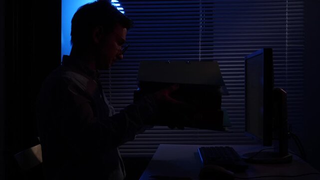 Man in silhouette working late at desk, reviewing documents and using a computer with cool blue light filtering through window blinds, focused and isolated at midnight