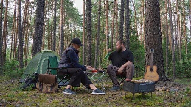 Young couple enjoying a picnic while sitting on chairs in a beautiful pine forest, with the woman slicing bread and the man taking vegetables from the table next to their tent and barbecue