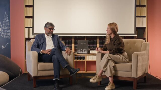 Handheld zoom out shot of male author talking about his book while man sitting in chairs on small stage with interviewer
