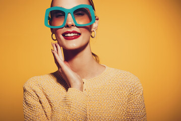 Smiling woman in oversized turquoise glasses and yellow sweater poses against orange background....