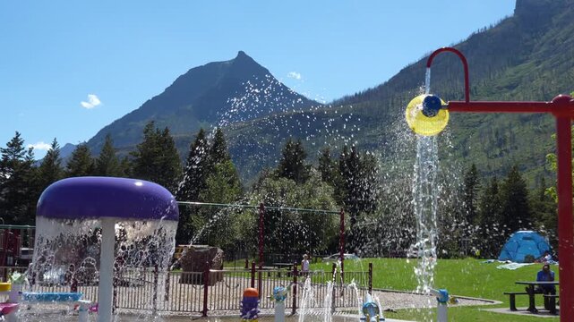 Bright splash pad with water features, a towering mountain background, and lush trees. Joyful, cool summer. Waterton, AB, Canada