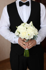 Groom in a vest and bow tie holds a bouquet of white roses before the wedding ceremony.