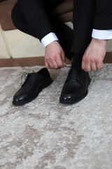 Groom ties black shoes while sitting on a sofa during wedding preparation.
