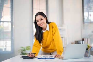 Asian young woman calculating financial data using a calculator and reviewing graphs on paper while...