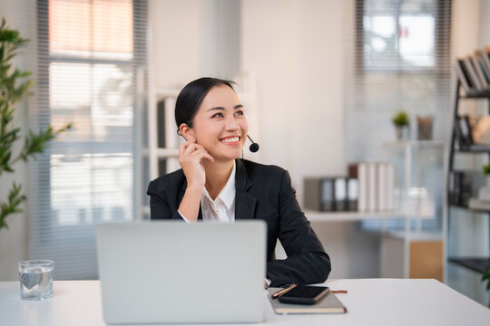 Asian business woman sitting at desk wearing a headset, smiling and looking up while providing excellent customer support in a modern office environment