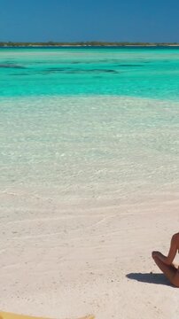 Aerial view of a young woman sitting on the sand of a pristine tropical beach, with a yellow kayak next to her and crystal clear turquoise water in the background