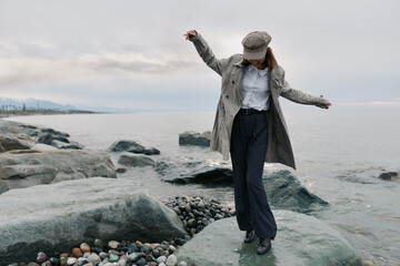 Obraz premium Woman in stylish coat and cap balances on rocky shore near calm sea under cloudy sky. Outdoor fashion, leisure, and nature concept with casual autumn outfit and relaxed mood.