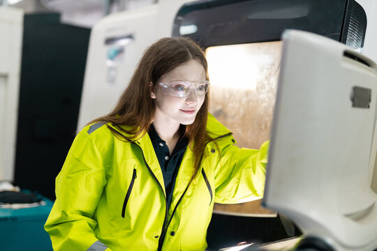 Female industrial engineer wearing safety jacket and protective goggles operating CNC machine in modern factory, focused on quality control, automation process, advanced manufacturing technology.