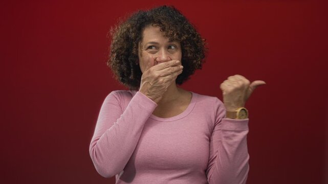 Woman of hispanic descent with curly hair wearing pink shirt covers mouth with hand and points thumb in red studio; surprise.