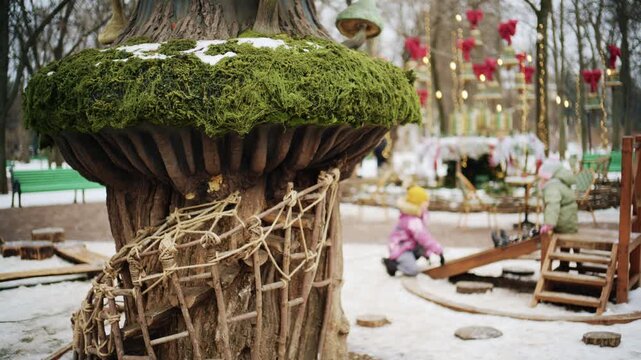 Winter park scene with children playing near a whimsical treehouse style structure decorated with moss and fairy tale elements