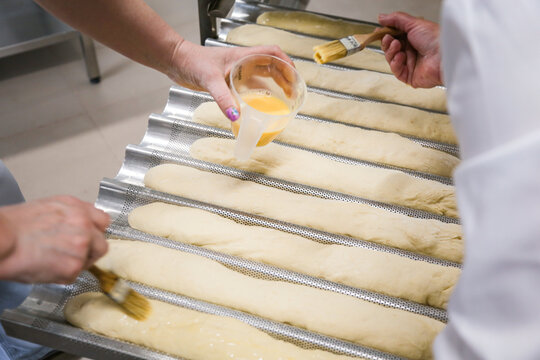 Women brushing baguettes with egg wash in a bakery kitchen before baking. Traditional bread preparation process in professional food production environment.