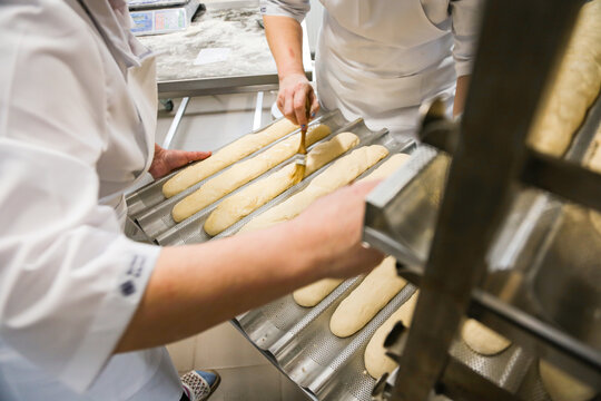 Women brushing baguettes with egg wash in a bakery kitchen before baking. Traditional bread preparation process in professional food production environment.