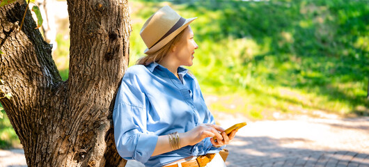 Smiling woman in hat stands near tree and holds smartphone. City travel and digital nomad lifestyle concept. © 5ph
