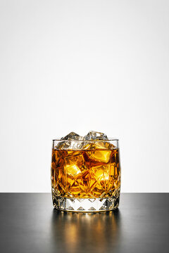 Close-up of a golden whiskey on the rocks in a faceted crystal rocks glass filled with large ice cubes, on a glossy black surface against a clean white background.