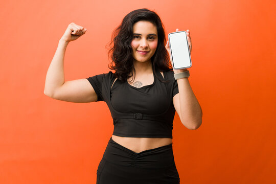 Empowered woman showing bicep and smartphone with blank screen, promoting fitness and health app