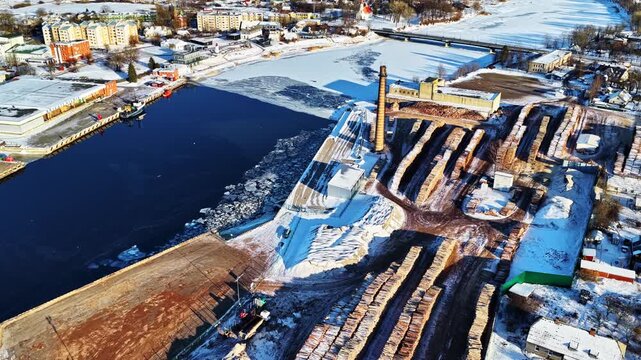 Salacgrīva port showcasing winter operations, logs stockpiled along the frozen river, and local town in aerial view on sunny winter day