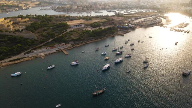 Cinematic aerial drone shot at sunset showcasing dozens of sailboats and yachts anchored in Malta's sparkling turquoise harbor, with coastal development and natural shoreline visible under golden ligh