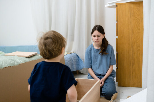 An older sister trying to assemble a wardrobe while her mischievous younger brother climbs inside the partially built structure, interfering playfully.