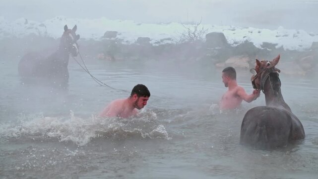 A man performs a backflip into a steaming natural hot spring while others bathe horses in winter. Snowy landscape, rising mist and dramatic action create a unique outdoor scene.