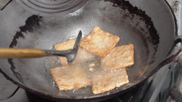 Hand holding a spatula flips fried tempeh in a pan. Close-up of nearly cooked tempeh.