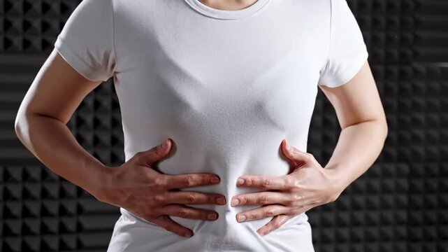 Asian woman in a white t-shirt gently holds her stomach with both hands, showing a gradual progression of her posture in a soundproof studio with textured walls