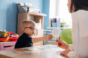 Preschool child with glasses interacts with teacher at a table, holding a card while educational...