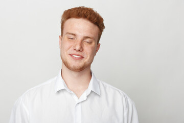 Obraz premium Young man with red hair and beard smiles gently with closed eyes wearing white shirt on light background. Concept of happiness, relaxation, and natural expression.