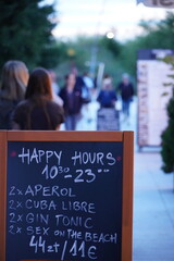 Menu z ofertą Happy Hour przed barem, ceny w złotówkach i euro, rozmyte postacie turystów w tle wieczorową porą. © bilcka