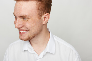 Fototapeta premium Smiling young man with red hair and freckles looks to the side wearing a white shirt. Studio portrait on a light background with natural expression and casual style.