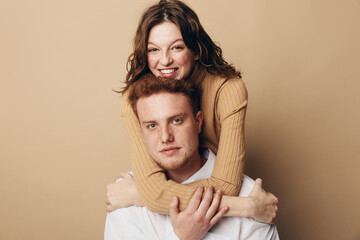 Fototapeta premium Smiling woman with curly hair hugs serious man from behind on beige background. Young couple shows affection and connection in casual clothing. Concept of love, relationship, and friendship.