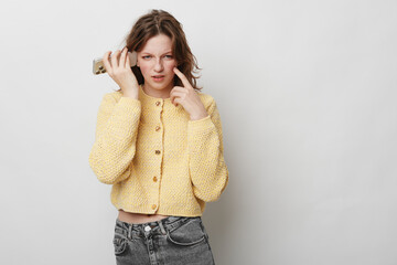 Confused young woman in yellow knitted sweater holds smartphone to ear and points to face,...