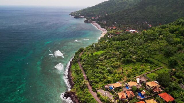 Drone aerial video captures the scenic shoreline of Bali Amed with transparent emerald water and a long rocky beach. A coastal village and tropical hills surround the calm ocean bay in Indonesia.