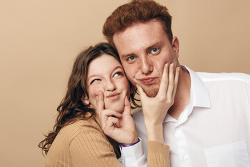 Fototapeta premium Young woman playfully pinches young man’s cheeks while both smile and pose against beige background. Concept of friendship, fun, affection, and casual lifestyle in studio portrait.
