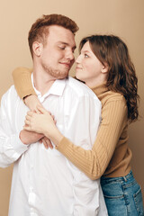 Fototapeta premium Young couple embraces tenderly, woman hugging man from behind with warm smile. Casual clothing and neutral background create intimate, loving, and affectionate mood in studio portrait.